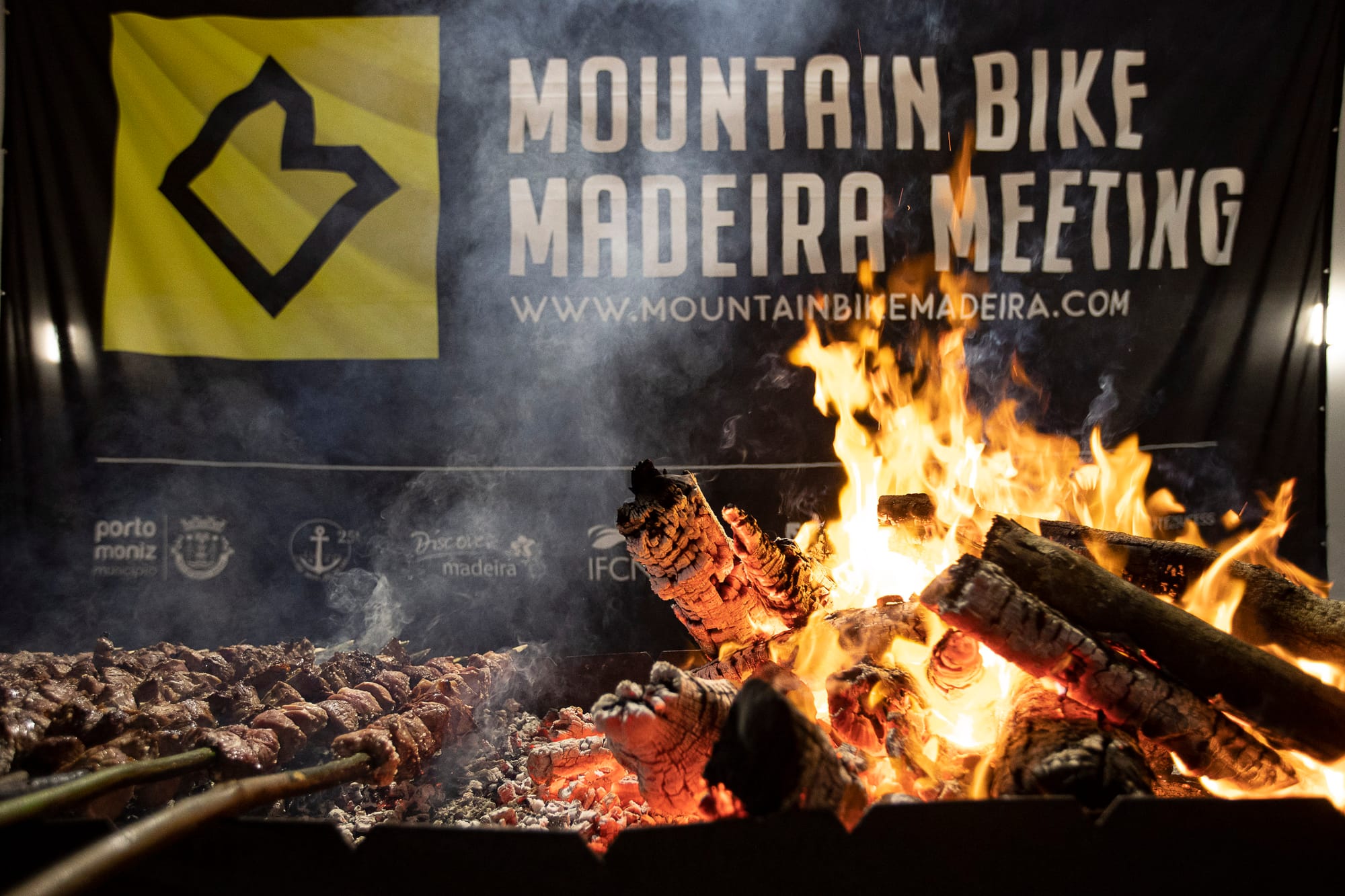 Espetada skewers over an open fire in front of the Mountain Bike Madeira Meeting banner